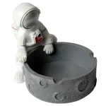 En ashtro-naut i keramik pryder overfladen med en glat, hvid finish, der understreger detaljerne i astronautens dragt. Hjelmen er blank med en reflekterende overflade og små, indgraverede detaljer. Rummet til aske er dybt og cirkulært, placeret i astronautens midte, mens figuren står på en solid, flad bund, der giver stabilitet.