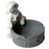 En ashtro-naut i keramik pryder overfladen med en glat, hvid finish, der understreger detaljerne i astronautens dragt. Hjelmen er blank med en reflekterende overflade og små, indgraverede detaljer. Rummet til aske er dybt og cirkulært, placeret i astronautens midte, mens figuren står på en solid, flad bund, der giver stabilitet.