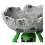 Et sæt af to askebægre i keramik præsenterer sig med en grøn alien-figur og en måneform. Alien-figuren har store, sorte øjne og en glat overflade, mens månen er dækket af fine detaljer og kraterlignende strukturer. Begge askebægre har en glansfuld finish, der fremhæver de levende farver og unikke former.