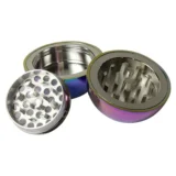 En kugleformet grinder med en diameter på 55 mm præsenterer sig i et glitrende regnbuefarvet design. Overfladen har en metalisk glans med en fin, struktureret tekstur. Den er opdelt i to halvkugler, hvor den øverste del har små, skarpe tænder til formaling. Der er en lille, diskret låsemekanisme i kanten, der sikrer lukning.