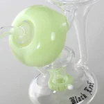 En sort, bladbølgende figur med glatte, afrundede kanter dominerer. Den har en dyb, ensartet overflade, der skaber en let refleksion. Materialet fremstår som plastisk med en mat finish. Under figuren ses en grøn, recycle-symbolet, der er indgraveret i en cirkulær form. Målene er kompakte, og elementerne er nøje centreret.