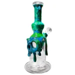 En keramikbong i sort og blå farve med en skulpturel uldne fåreskalle formet hoved. Skallen har detaljerede teksturer og kurver, der fremhæver dybderne i materialet. Bongen har en cylindrisk form med en transparent glaskolbe, der tydeligt viser væsken indeni. Den har en bred fod for stabilitet og en buet hals, der afsluttes med en åbning.