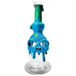 En sort får skull med detaljeret tekstur og dybe fordybninger pryder toppen af en blå bong. Bongen har en glat, blank overflade med en cylinderformet krop, der afsluttes med en bred åbning. Farven skifter fra dyb blå til en lysere nuance mod bunden. Der er en lille, transparent perle ved siden af skull-designet.