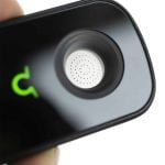En kompakt vaporizer med en slank, sort krop og en mat overflade. Den har en beskyttende låge, der dækker en glas vandkammer. På fronten findes en digital skærm med tydelige, hvide tal og symboler. En metalknap til betjening er placeret på siden. En fleksibel slange til inhalation er koblet til en åbning i enheden.
