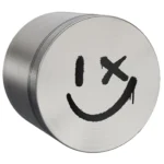 En sølvfarvet metalgrind med en diameter på 63 mm præsenterer et glat, reflekterende overflade. Grindens design inkluderer et smilende ansigt i midten, der er præcist indgraveret med mørke detaljer, hvilket skaber kontrast til den lyse baggrund. Kanten er let bølget, og der er små ventilationshuller, som tilføjer tekstur og detaljer til det samlede udseende.