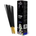 En pakke indeholdende røgelse med en dyb blå farve pryder forsiden. Et stiliseret cannabisblad er placeret centralt, omgivet af subtile mønstre og tekst i hvid og guld. Teksten angiver produktnavnet "Blueberry Haze" i en moderne skrifttype. Baggrunden er en let tekstureret sort, der fremhæver de farverige elementer. Formen er rektangulær med afrundede hjørner.