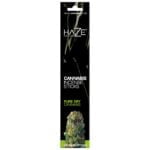 En transparent beholder indeholder tørret cannabis, der præsenterer en rig grøn farve med nuancer af brun og gylden. Planten har en fyldig, klæbrig tekstur og små, tydelige trichomer, der glitrer let. Beholderens låg er af klart plastmateriale, og etiketten er hvid med sort tekst, der angiver produktinformation.