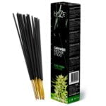 En hvid emballage prydes af en stiliseret grøn plante, der ligner cannabis, med en let skyggeeffekt. Øverste del af pakken viser tydelig tekst i sort, der angiver produktets navn. Bunden har en baggrund i en lys grøn nuance, hvilket skaber en kontrast til den hvide overflade. Det hele har en glat, mat tekstur.