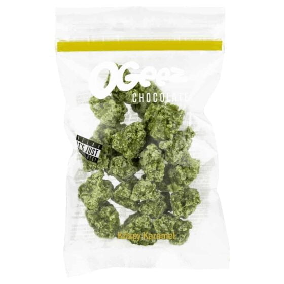 OgeeZ Krispy Karamel præsenterer sig i en papemballage med en dominerende lysebrun farve, prydet med gyldne og hvide detaljer. Fronten viser produktnavnet i store, tydelige bogstaver samt en illustration af en sprød snack. Emballagens tekstur fremstår glat, mens kanten er let buet. Baggrunden har et subtilt mønster, der tilføjer visuel dybde.