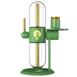 Stundenglass Gravity Hookah præsenterer en elegant, glasklar beholder med en afrundet form, der strækker sig op til en smal hals. Farven skifter fra klar til en dyb grøn, og refleksionerne fra glasset skaber en glitrende effekt. Den metalliske top med en børstet finish har flere tilslutninger og en enkelt, sort slange, der bølger ud fra siden.