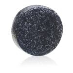 En solid shampoo i en rund, flad form præsenteres med en glat, beige overflade. Massen har en let skinnende tekstur, som reflekterer lyset blidt. På toppen er der prægede detaljer med et mønster, der giver et rustikt udtryk. Vægten er 60 gram, og der ses ingen synlige indpakninger eller etiketter.