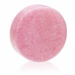 En rund, flad skive af solid shampoo med en lys, pastelrosa farve. Overfladen er glat og skinnende, med en let struktur, der giver en følelse af blødhed. Der er indprægede mønstre i overfladen, og kanterne er let afrundede. Vægten er angivet til 60 gram, og produktet er placeret på en neutral baggrund, der fremhæver dets farve.