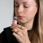 En rund, flad beholder i klart glas med en tynd, hvid metal låg. Indholdet består af grove, hvide krystaller, der ligner salt, og har en let glitrende overflade. Beholderens side er prydet med en enkelt, elegant etikette, der indeholder tekst i en simpel sort skrifttype. Låget har en let børstet finish, hvilket tilføjer en subtil tekstur.