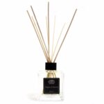En klar glasflaske med en stram, cylindrisk form indeholder lysebrun væske. Flasken har en sort, tætsiddende korkprop, der holder aromaen indkapslet. Ved siden af flasken står en sætning af elegante, smalle pinde i lys træ, der er justeret til at stikke opad. Baggrunden er lys og neutral, hvilket fremhæver flaskens farver og materialer.