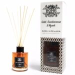 En elegant diffuser med en cylindrisk glasbeholder i klar glas, prydet med en skinnende guldfarvet metalring øverst. Indholdet er en gylden væske, der reflekterer lyset. Stifterne, der stikker op fra beholderen, er brune og slanke. Etiket på glasset er diskret med tekst, der angiver duftnoter af frankincense og myrra.