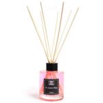 En elegant diffuser i glas præsenterer sig med en klar, cylindrisk form. Den indeholder en transparent væske, der let reflekterer lys. Øverst er der en smuk, hvid korkprop, som komplementerer det minimalistiske design. Syv lange, smalle rør af naturlige materialer stikker op fra proppen, hvilket tilføjer en tekstur og et organisk udtryk. Farverne er bløde og neutrale.