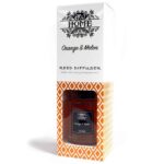 En rektangulær diffuser i glas med en klar, glat overflade præsenterer en orange væske indeni. Diffuseren er fyldt med rørformede, naturlige stænger i lys træfarve, der stikker op fra væsken. Et simpelt, hvidt etiket med sort tekst pryder fronten. Baggrunden er neutral og blød, hvilket fremhæver diffuserens farver og materialer.