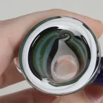 En skål med en dyb, rund form præsenterer sig med en glat, sort glasoverflade. Mørkegrønne spiralmønstre snor sig elegant gennem skålens indvendige væg, hvilket skaber en kontrast til den sorte baggrund. Kanten er let bølget, og skålen har en poleret, reflekterende finish, der tilføjer dybde og dimension til designet.
