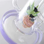 En høj, slank bong i sort glas prydet med detaljer i lilla. Den har en buet form og en bred, stabil fod. Overfladen er glat, mens den lilla farve skaber en kontrast ved slangen og toppen. En perkolator ses indeni, hvilket tilføjer et komplekst design. Bongen har en indbygget skål og en tæt passform for sikker anvendelse.