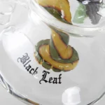 En sort olie-rør med en skulpturel skildpaddeform, fremstillet af glas med en glat overflade. Røret har en perkolator, der skaber bobler i væsken, hvilket giver en interessant visuel effekt. Det har runde, bløde former med en let buet hals og en flad bund, der sikrer stabilitet. Nogle detaljer i klar glas fremhæver dets design.