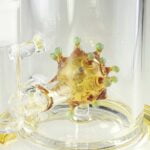 En gul glas dabbing rig med en karakteristisk virusformet struktur pryder midten. Den er 24 cm høj, og dens overflade er glat med en let gennemsigtig finish. Rigen har en buet hals og en bred base, der sikrer stabilitet. Der er en klar perkolator nederst, som tilføjer dybde til designet. Det samlede udtryk er organisk og kreativt.