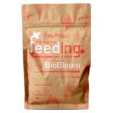 En papbeholder i grøn farve prydes af en detaljeret etiket med hvid og sort tekst. Etiketten fremhæver produktnavnet "BioBLOOM" i stor skrifttype. Beholderens låg er skrueformet med en glat overflade. Under etiketten er der angivet vægten "1 kg" samt instruktioner og ingredienser i mindre skrifttype. Materialet fremstår mat og robust.