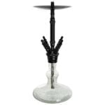 En cylindrisk lampe med en mat sort overflade pryder midten af kompositionen. Lampen har en let buet form og er fremstillet af metal. Øverst er der en rund, oplyst diffusor, der udsender et blødt, varmt lys. Nederst er der en smal, flad fod, der stabiliserer lampens opretstående position, mens ledningen elegant er placeret langs siden.