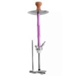 En stor, firkantet paraply med en dyb lilla farve dominerer scenen. Stoffet er glat og har en let skinnende overflade, hvilket skaber en elegant effekt. Paraplyen er monteret på en solid, sølvfarvet metalstang med en robust base, der sikrer stabilitet. Kanten er afsluttet med en bøjelig kant, der giver en blødere silhuet.