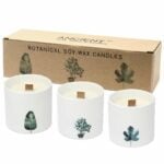 Store botaniske stearinlys pryder en hvid overflade, omkranset af grønne planter. Lysene har en blød, rund form med en glat overflade, prydet med naturlige mønstre og farver, der minder om planter og blomster. De er placeret i en uformel gruppe, hvilket skaber en harmonisk og indbydende atmosfære med en naturlig æstetik.
