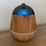 En aromadiffuser i en elegant, cylindrisk form er fremstillet af let træmateriale med en glat, naturlig finish. Toppen har en flad, rund åbning, hvor damp og duft frigives. En USB-port er placeret i bunden, og en lille knap til aktivering er synlig. Farven er varm og lys, hvilket giver et rustikt udseende.