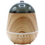 En aromadiffuser i trædesign præsenterer en cylindrisk form med en glat, lys finish. Toppen er let afrundet og inkluderer en knap til betjening. En USB-port er synlig på siden, og der er små ventilationshuller ved bunden. Diffuseren er placeret på en flad overflade, omgivet af en minimalistisk indretning med naturlige elementer.