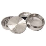 En metalgrinder i rustfrit stål med en diameter på 63 mm. Den har en glat, poleret overflade, der reflekterer lys. Grinderens top har et skridsikkert greb i en mat finish, mens det indre er udstyret med skarpe tænder til effektiv formaling. Kanten er let afrundet, og der er små, fine detaljer ved samlingerne.