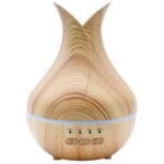 En USB-diffuser i en elegant, cylindrisk form præsenterer sig i et naturligt trælook. Overfladen har en glat, mat finish med subtile åretegninger, der fremhæver træets struktur. En gennemsigtig, rund top med en let buet form afslutter designet. En lille knap og en USB-port er anbragt på siden, hvilket giver en minimalistisk og moderne æstetik.