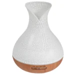 En aromadiffuser i en rund form med en mat hvid overflade dominerer designet. Den har en træfarvet bund, der giver en naturlig kontrast. Øverst er der et gennemsigtigt låg, som afslører en indvendig vandbeholder. Diffuseren har en USB-port til strømforsyning og tydelige knapper til betjening. Mønstret på overfladen tilføjer en subtil tekstur.