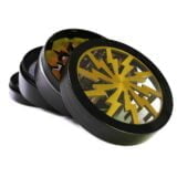 En metal grinder med en gylden overflade pryder midten af beskrivelsen. Den har en cylindrisk form med en diameter på 50 mm. Overfladen er glat og skinnende, og der er fine riller langs kanten for bedre greb. Grinderen har små, skarpe tænder indeni, der er designet til at male materialer. Der er også en lille rund åbning på toppen, muligvis til påfyldning.