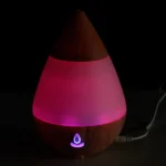En dråbeformet aroma diffuser i lys træfinish med en glat overflade. Den har en rund bund og en tilspidset top, der giver et elegant udtryk. På forsiden er der en lille kontrolknap, der er indlejret i træet. Diffuseren har en diskret USB-port til tilslutning. Det samlede design udstråler naturlighed og enkelhed.
