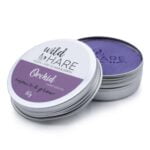 En solid shampoo i en rund, flad form med en glat, mat overflade. Farven er en blid lavendel, der fremkalder associationer til orkidéer. Produktet er indpakket i en minimalistisk, hvid papæske med grøn tekst, der indeholder informationer om ingredienser og anvendelse. Æsken har en simpel, naturlig æstetik uden unødvendige dekorationer.