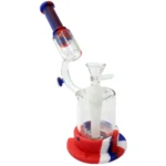En 21 cm høj bong i gennemsigtig silikone med en hexagonal form, der præsenterer en klar, farverig struktur. Overfladen har en glat tekstur, mens de runde kanter giver en blød profil. Bongen inkluderer en skål og en rørformet hals, som begge er designet til at matche den overordnede æstetik med en farverig og moderne finish.