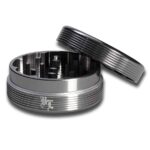 En sort krydderikværn i metal med en diameter på 50 mm præsenterer sig med bølgede blade, der skaber et struktureret mønster på overfladen. Kværnen har en glat, mat finish, der kontrasterer med den blanke inderside. Der er en lille skrue i midten, der bruges til at justere knivene, og selve kværnen har en rund form med en skruelukning.