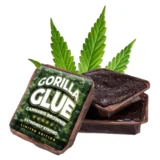 En fyldig, brun brownie præsenteres med en glat, let skinnende overflade, der indikerer en rig chokoladefarve. Kanten er let sprød, mens midten fremstår blød og saftig. Små stykker nødder er synlige og tilføjer struktur. Den samlede form er firkantet, og bagværket ligger på en neutral, lys overflade, der fremhæver farven.