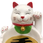 Et askebæger i klar glas prydet med en dekorativ lucky cat figur i keramisk materiale. Figuren er malet i en kombination af hvid, sort og gylden farve, med detaljerede ansigtstræk og en udstrakt pote. Askebægeret har en cirkulær form med glatte kanter, og bunden er let skrånet for stabilitet. Det klare glas giver en elegant og moderne fremtoning.