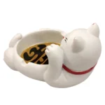 Et askebæger i porcelæn præsenterer sig med en hvid overflade, dekoreret med en farverig kattefigur, der sidder med en oprejst pote. Katten har sorte, røde og gule detaljer, herunder et smilende ansigt og gyldne accenter. Bunden er flad, mens kanten er let hævet, hvilket skaber en skarp kontrast til den glatte tekstur.