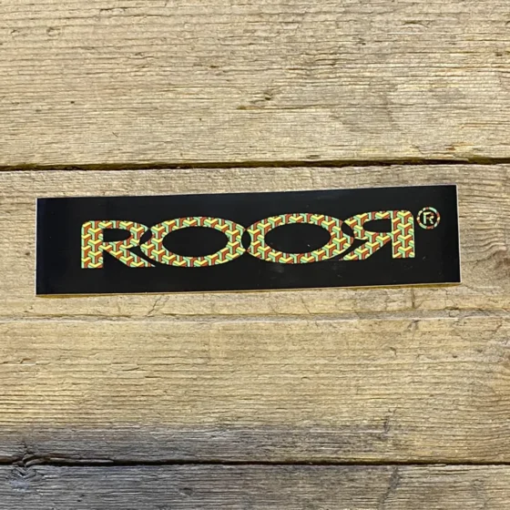 Rør-logoet fremtræder i en kontrastfuld kombination af sort og gul. Det sortfarvede område udgør baggrunden, mens det gule logo er designet med skarpe kanter og en tydelig skrifttype. Bogstaverne er store og klare, hvilket giver en markant visuel effekt. Materialet ser glat ud, hvilket indikerer en plastisk tekstur, ideel til klistermærker.