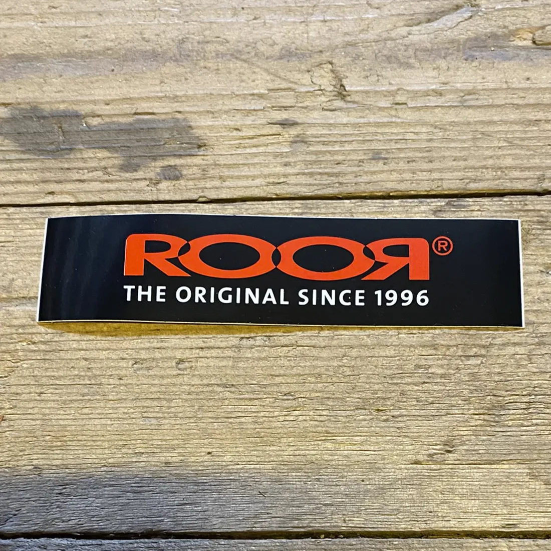 Et klistermærke i rektangulær form præsenterer et logo med en kombination af sort og rød farve. Logoet indeholder grafiske elementer og tekst i en moderne skrifttype. Den sorte baggrund fremhæver det røde design, hvilket skaber en kontrast. Klistermærket har en glat overflade med en let refleksion, der indikerer en plastisk kvalitet.