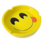 Et askebæger i klart glas prydes af en glad smiley i gule og sorte farver. Smileyen har runde øjne og en bred mund, der udstråler glæde. Bægeret har en glat overflade med en let buet form og en solid base, hvilket giver stabilitet. Det gennemsigtige glas skaber en elegant fremtoning, der understreger smileyens farver.