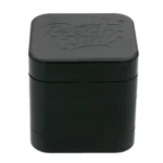 En sort grinder med en firkantet form og en diameter på 50 mm. Overfladen har en mat finish, der giver en elegant tekstur. Kanten er let afrundet, og der er små, præcise tænder indeni til at male. Grinderen har en enkelt, snæver åbning øverst og en slank bund med en tydelig skillelinje, der adskiller de to dele.