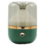 En aroma diffuser præsenterer sig i en kombination af grønne og bronzefarvede nuancer. Den har en rund, glat form med en mat overflade og en let struktur. Øverst er der en åbning med en diskret, rund knap. Diffuseren har en belysningseffekt, der skaber en blød glød, og dens bund er stabil og flad.