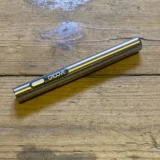 En sølvfarvet batteri med en slank, cylindrisk form pryder overfladen. Det har en glat metaltekstur og en mat finish. Øverst er der en rund kontakt med et blåt lys, og under er en skruegevind til tilslutning. En lille, indgraveret logo findes på siden, mens bunden har en skridsikker overflade.