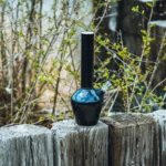 En 33 cm høj bong i glat, glansfuld blå farve med en blank overflade, fremstillet af rustfrit stål. Den har en rund base, der gradvist snævrer ind mod en cylindrisk hals. En krum rørføring fører til en mundstykke, og der er en indsat skål til rygning. Det samlede design er minimalistisk med en moderne æstetik.