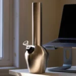 En sølvfarvet bong i rustfrit stål med en højde på 33 cm. Den har en glat, skinnende overflade, der reflekterer lys. Bongen er designet med en buet hals og en bred base, hvilket giver stabilitet. Der er en perkolator i bunden og en rørformet mundstykke, der afsluttes med en rund åbning. Det minimalistiske design fremhæver dets funktionalitet.